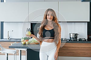 Holds apple in hand. Young european woman is indoors at kitchen indoors with healthy food