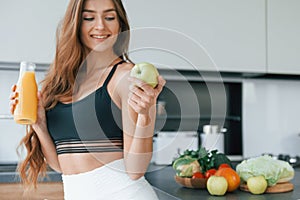 Holds apple in hand. Young european woman is indoors at kitchen indoors with healthy food