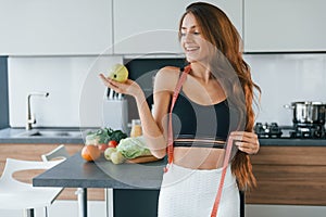 Holds apple in hand. Young european woman is indoors at kitchen indoors with healthy food