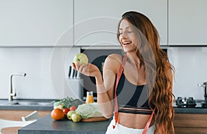 Holds apple in hand. Young european woman is indoors at kitchen indoors with healthy food