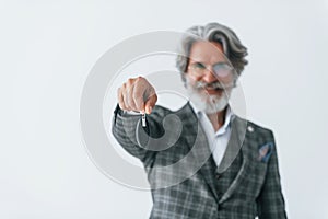 Holds car keys in hand. Senior stylish modern man with grey hair and beard indoors