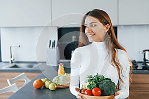 Holds plate with vegetables. Young european woman is indoors at kitchen indoors with healthy food