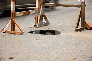 Hole in asphalt on automotive roadway street. Pit in road surface, is surrounded by building fence construction with warning tape