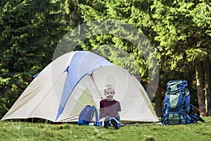 Holiday camping.Happy young boy sitting in front of a tent near backpacks taking rest after hiking in the forest.Traveling and tou