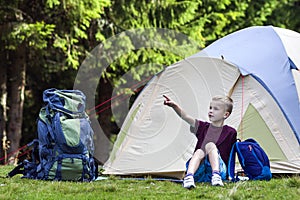 Holiday camping. Young boy sitting in front of a tent near backpacks taking rest after hiking in the forest shows something in the