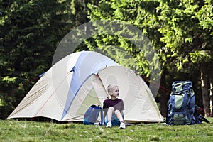 Holiday camping. Young boy sitting in front of a tent near backpacks taking rest after hiking in the forest shows something in th