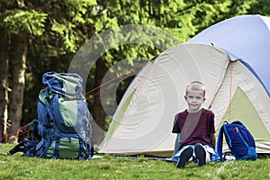 Holiday camping. Young boy sitting in front of a tent near backpacks taking rest after hiking in the forest shows something in th