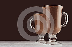 Holiday chocolate drinks in elegant transparent goblets on dark brown background, white wood board, copy space.