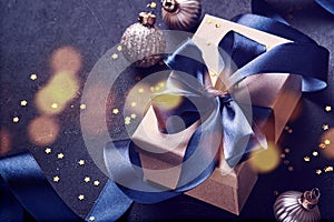 Holiday gift box or present with blue ribbon, golden confetti, baubles and light blur bokeh on black background. Christmas greetin