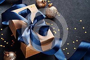 Holiday gift box or present with blue ribbon, golden confetti and gold baubles on black background. Magic christmas greeting card.