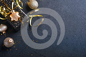 Holiday gift box or present with ribbon, golden confetti and gold baubles on black background. Magic christmas greeting card.