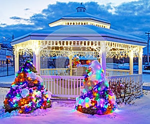 Holiday lights at night on white gazebo with snow on Christmas trees at night