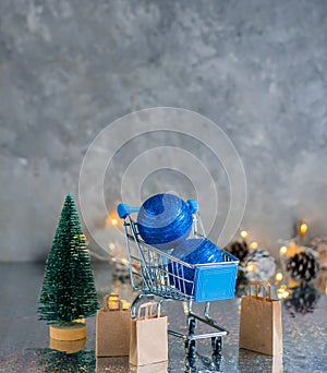 Holiday shopping. Shopping cart with blue shiny balls, Christmas tree and paper bags on a gray background with garland and lights