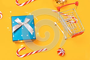 Holiday shopping. A toy shopping cart and a gift box. Yellow background with candys and christmas ball. Close up. Top view.