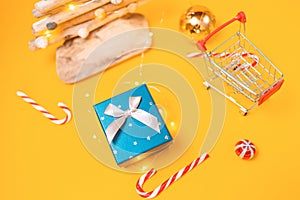 Holiday shopping. A toy shopping cart, gift box, and a craft wooden Christmas tree. Yellow background with candys and christmas