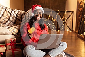 Holiday shopping. Young black woman in Santa hat using credit card and laptop, ordering Xmas gifts online from home