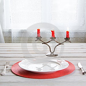 Holiday table place setting with white dishware, cutlery, silverware and red decorations on white wooden board in ligth interior n