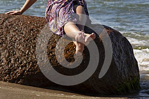 The holiday vacation. Woman legs close-up of a girl sitting on a large rock on the seashore, waves and splashes of water at her