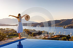 A holiday woman in a dress stands at the swimming pool and enjoys the summer sunset