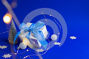 Holiday xmas background. White gift with blue ribbon, winter tree, Snowflakes and Silver balls in Christmas composition on blue