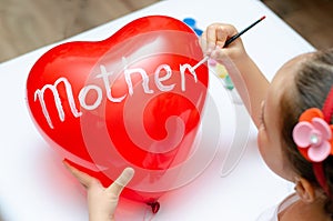 For the holiday, a 5-year-old girl writes the word mom with white paint on a heart-shaped balloon. Blurry, defocused, selective