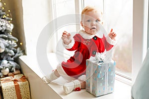 Holidays and childhood concept - sweet baby girl in santa costume sitting with xmas gift box at the window near christmas tree