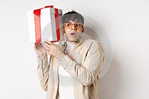 Holidays concept. Funny young man in glasses shaking gift box, listening what inside present and gasping amazed