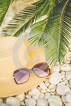 Holidays concept layout. Straw hat on the pubble beach under palm tree leaves. Vacation accesories