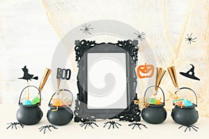 Holidays image of Halloween. Witcher cauldron, broom, candies and spiders next to empty photo frame for mockup over wooden table.