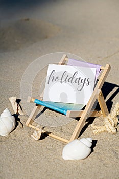 HOLIDAYS text on paper greeting card on background of beach chair lounge summer vacation decor. Sandy beach sun. Holiday