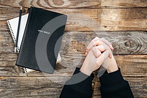 Holy Bible and male hands folded in prayer on a wooden background, top view.