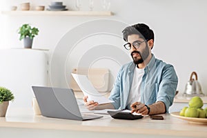 Home accounting, taxes. Happy young arab man in white minimalist kitchen interior, with computer and calculator