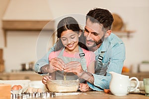 Home bakery concept. Happy father adding egg to cookies dough, helping and learning daughter how to cook