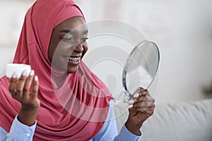 Home beauty treatment. Black muslim lady in hijab holding moisturising cream jar photo