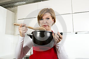 Home cook woman at kitchen holding cooking pot and spoon tasting soup in a funny disgusting bad taste face