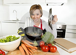 Home cook woman in red apron at domestic kitchen holding cooking pot with hot soup smelling vegetable stew