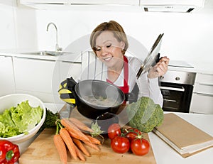 Home cook woman in red apron at domestic kitchen holding cooking pot with hot soup smelling vegetable stew