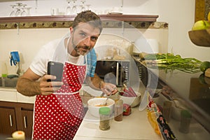 Home cook at work ! young happy and attractive home cook man in red apron following online recipe on internet mobile phone cooking