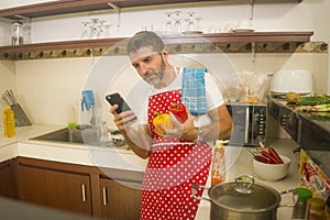 Home cook at work ! young happy and attractive home cook man in red apron following online recipe on internet mobile phone cooking