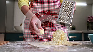 Home cooking of food. A woman in an apron grates cheese on a grater, and then shows a mound of grated product. Hands close up