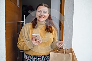 Home delivery. Woman with smartphone opening house entrance door while meeting courier with online order, postwoman