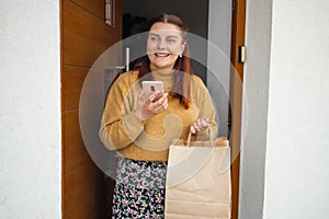 Home delivery. Woman with smartphone opening house entrance door while meeting courier with online order, postwoman