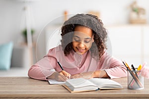 Home education concept. Cute african american schoolgirl doing homework and smiling, sitting at table with book