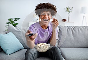 Home entertainments. Excited African American teenager with TV controller enjoying movie and eating popcorn indoors