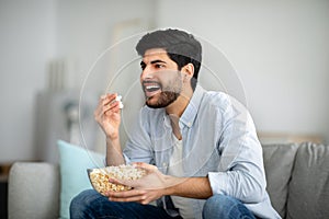 Home entertainments. Excited arab guy eating popcorn and watching TV, spending free time at home, free space