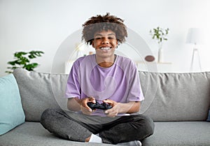 Home entertainments. Happy black teenager with joystick playing online computer games in sofa indoors, full length