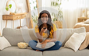 Home entertainments. Smiling black woman with remote control eating popcorn and watching TV on sofa in living room