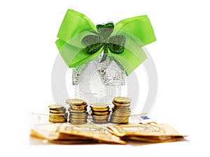 Home with glitter on stack of coins and banknotes and green bow with shamrock on white background. High value cost of property in