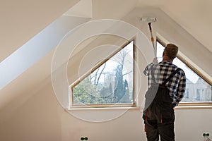 Home improvement concept, handyman painting a wall with a white paint near roof window in attic