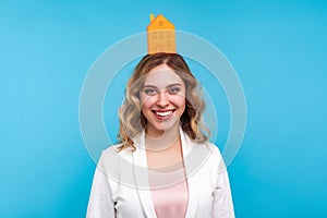 Home insurance. Portrait of positive woman standing with small paper house on her head and smiling broadly. blue background